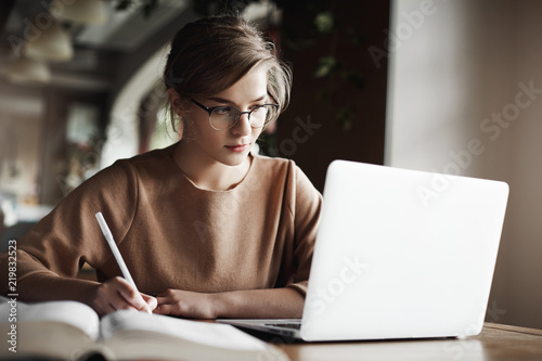 Creative good-looking european female with fair hair in trendy glasses, making notes while looking at laptop screen, working or preparing for business meeting, being focused and hardworking photo