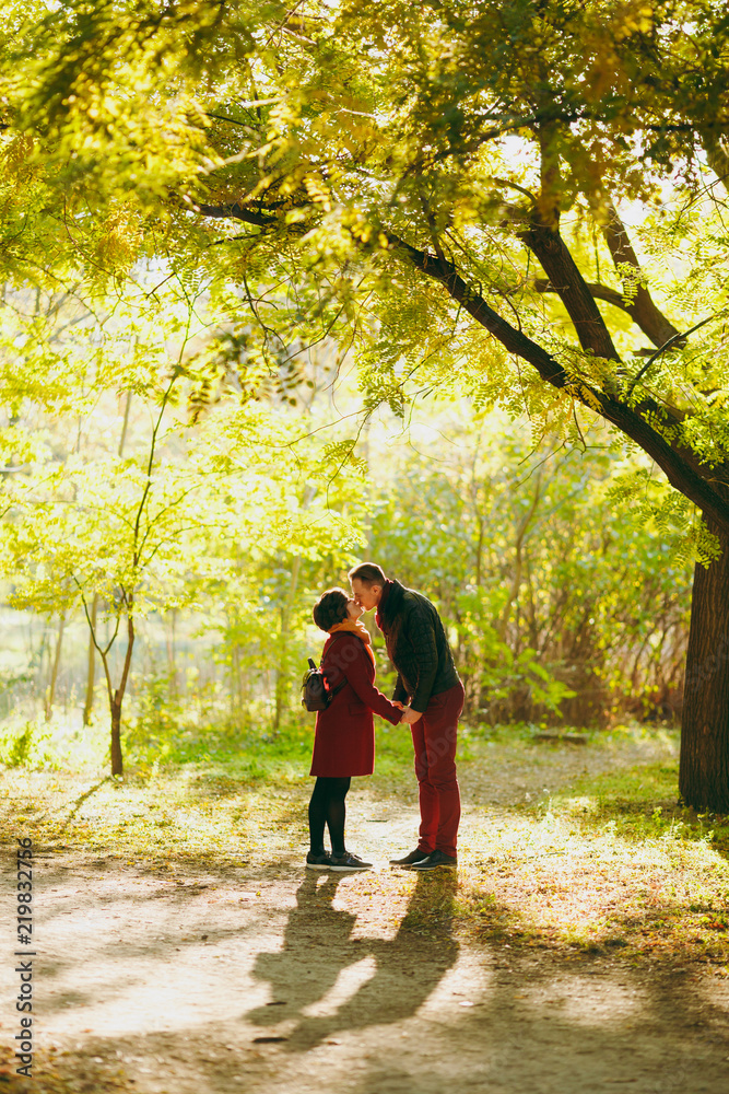 Young beautiful couple in love woman with backpack, man in casual warm clothes holding hands kissing in autumn city park outdoors in sunny weather. Love relationship family people lifestyle concept.