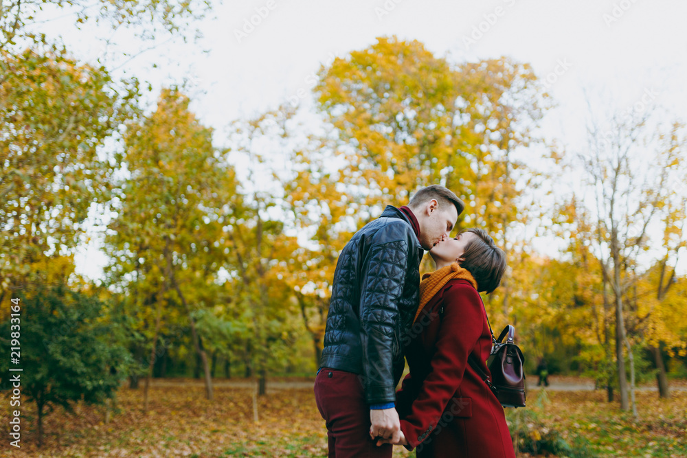 Young romantic couple in love woman and man in warm clothes holding hands kissing with closed eyes standing in autumn city park outdoors on trees background. Love relationship family people lifestyle.