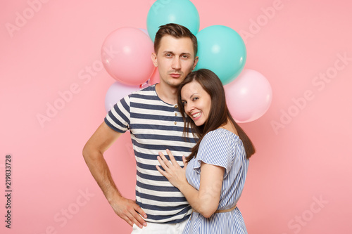 Portrait of young happy embracing couple in love. Woman lean on one's man chest celebrating birthday holiday party on pastel pink background with colorful air balloons. People sincere emotions concept