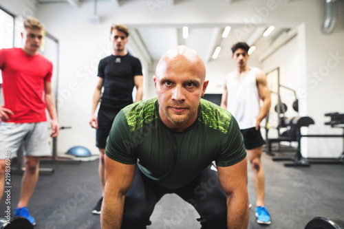 Fit young men looking at a personal trainer in gym lifting barbell.