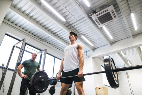 Fit young man with a personal trainer in gym working out, lifting barbell.