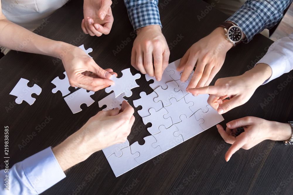 Top close up view of diverse employees assembling jigsaw puzzle trying to find best business solution, workers engaged in teambuilding practicing unity and support. Concept of successful cooperation Top close up view of diverse employees assembling jigsaw puzzle trying to find best business solution, workers engaged in teambuilding practicing unity and support. Concept of successful cooperation