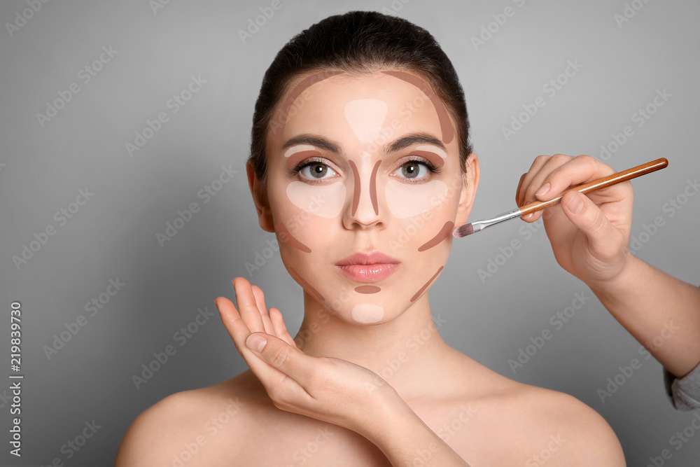 Naklejka premium Wizaż artysta stosuje makeup na kobiety twarzy przeciw popielatemu tłu. Profesjonalne produkty kosmetyczne