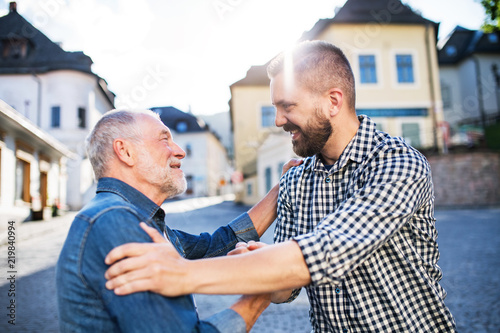 An adult hipster son with senior father on a walk in town, greeting.