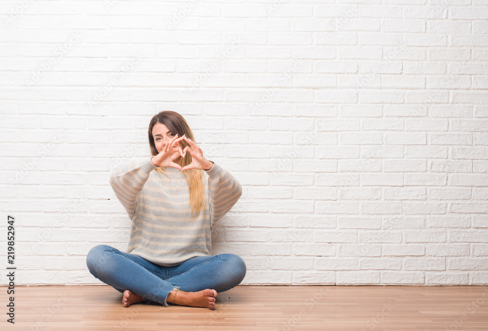 Young adult woman sitting on the floor over white brick wall at home smiling in love showing heart symbol and shape with hands. Romantic concept. Young adult woman sitting on the floor over white brick wall at home smiling in love showing heart symbol and shape with hands. Romantic concept.