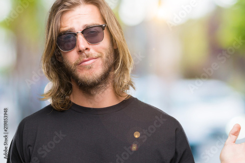 Young handsome man with long hair wearing sunglasses over isolated background clueless and confused expression with arms and hands raised. Doubt concept.