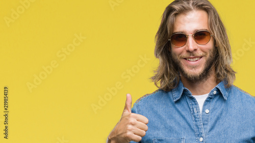 Young handsome man with long hair wearing sunglasses over isolated background doing happy thumbs up gesture with hand. Approving expression looking at the camera with showing success.