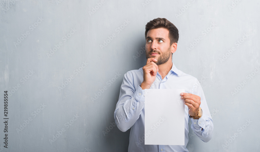 Handsome young man over grey grunge wall holding blank paper sheet contract serious face thinking about question, very confused idea Handsome young man over grey grunge wall holding blank paper sheet contract serious face thinking about question, very confused idea