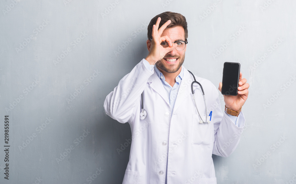 Handsome young doctor man over grey grunge wall holding smartphone with happy face smiling doing ok sign with hand on eye looking through fingers Handsome young doctor man over grey grunge wall holding smartphone with happy face smiling doing ok sign with hand on eye looking through fingers
