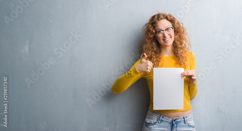 Young redhead woman over grey grunge wall holding blank paper sheet happy with big smile doing ok sign, thumb up with fingers, excellent sign photo