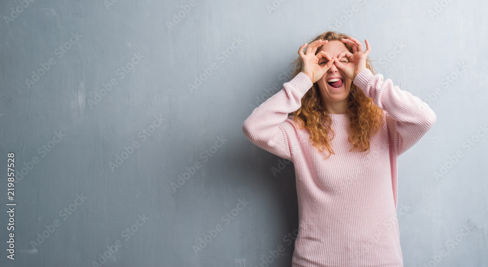 Young redhead woman over grey grunge wall wearing pink sweater doing ok gesture like binoculars sticking tongue out, eyes looking through fingers. Crazy expression.