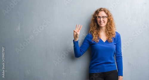 Young redhead woman over grey grunge wall showing and pointing up with fingers number three while smiling confident and happy.