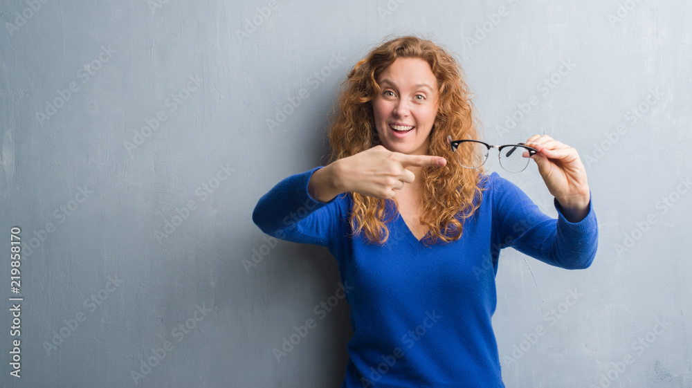 Young redhead woman over grey grunge wall holding optical glasses very happy pointing with hand and finger Young redhead woman over grey grunge wall holding optical glasses very happy pointing with hand and finger