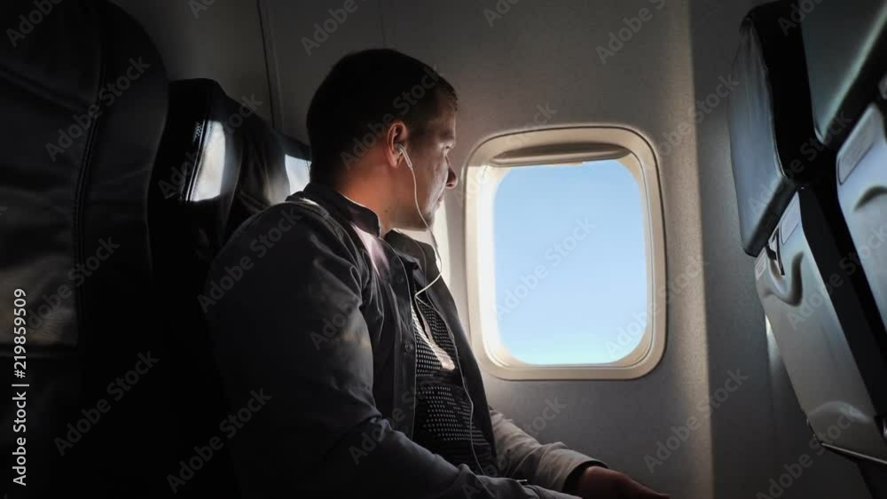The young man looks out the window of the plane in the evening. He listens to the headphones and rests.