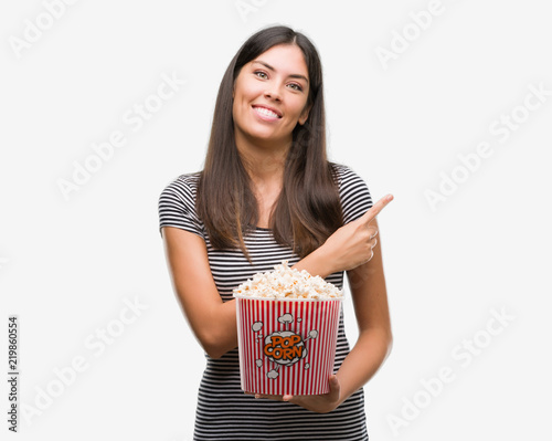 Young beautiful hispanic eating popcorn very happy pointing with hand and finger to the side