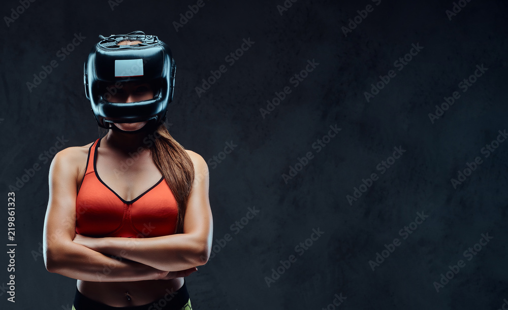 Sportive woman in sports bra wearing a protective helmet, posing with crossed arms and looking at a camera. Isolated on dark textured background.