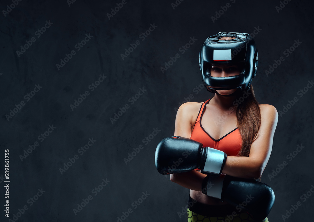 Sportive woman in sports bra wearing a protective helmet and boxing gloves, posing in a studio. Isolated on a dark textured background.