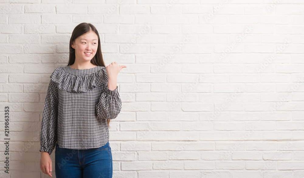 Young Chinese woman over brick wall pointing and showing with thumb up to the side with happy face smiling Young Chinese woman over brick wall pointing and showing with thumb up to the side with happy face smiling