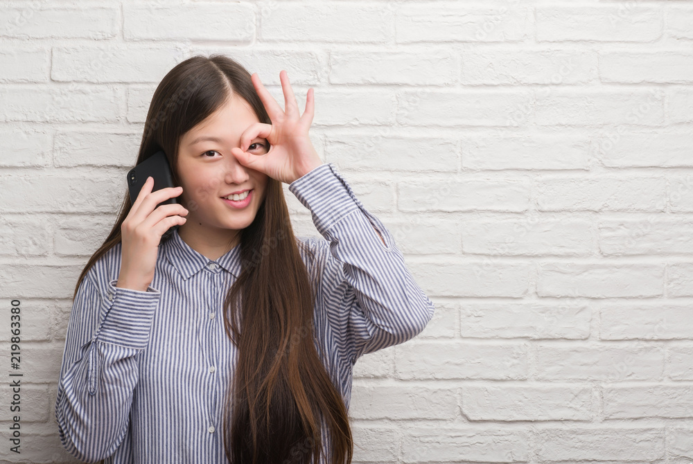 Young Chinese woman over brick wall talking on the phone with happy face smiling doing ok sign with hand on eye looking through fingers Young Chinese woman over brick wall talking on the phone with happy face smiling doing ok sign with hand on eye looking through fingers