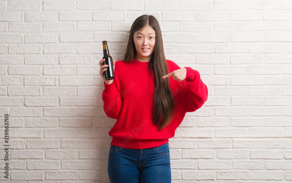 Young Chinese woman over brick wall drinking beer with surprise face pointing finger to himself Young Chinese woman over brick wall drinking beer with surprise face pointing finger to himself