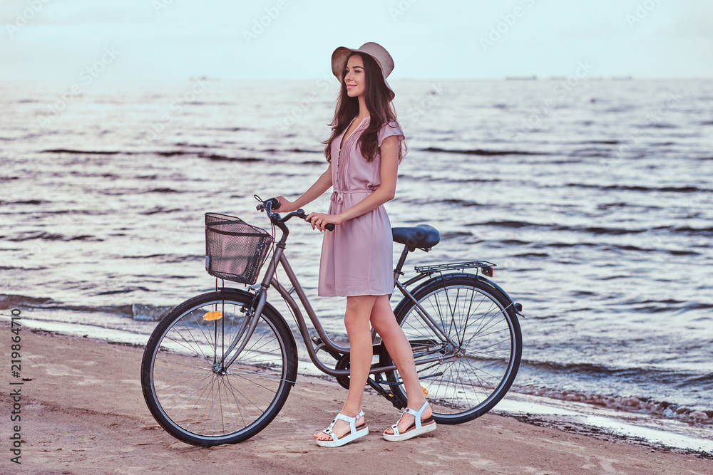 Fototapeta premium Happy sensual girl in hat wearing dress walks with her bicycle on the beach against amazing seaside background.