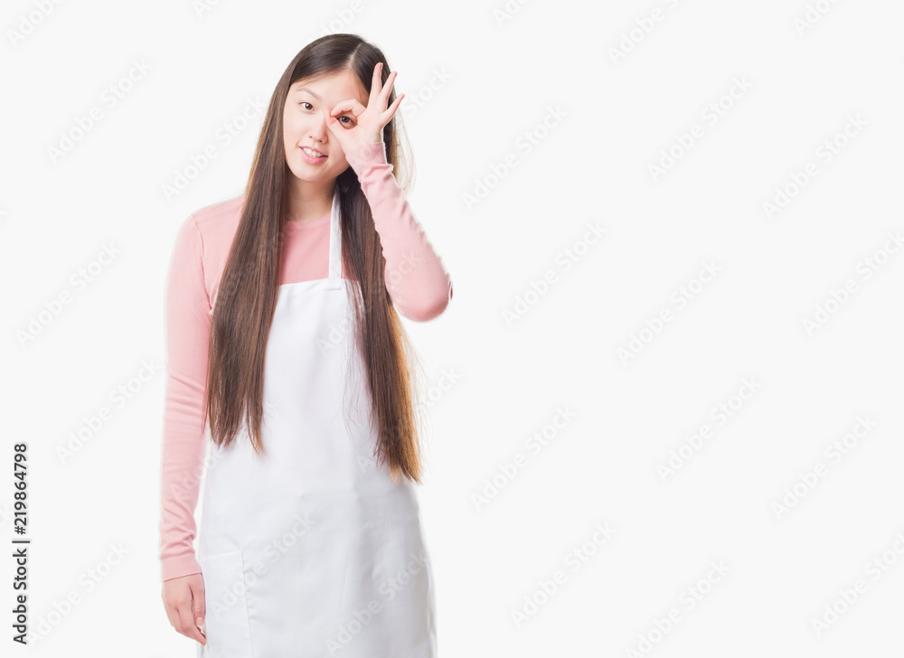 Young Chinese shop owner woman over isolated background wearing white apron with happy face smiling doing ok sign with hand on eye looking through fingers Young Chinese shop owner woman over isolated background wearing white apron with happy face smiling doing ok sign with hand on eye looking through fingers