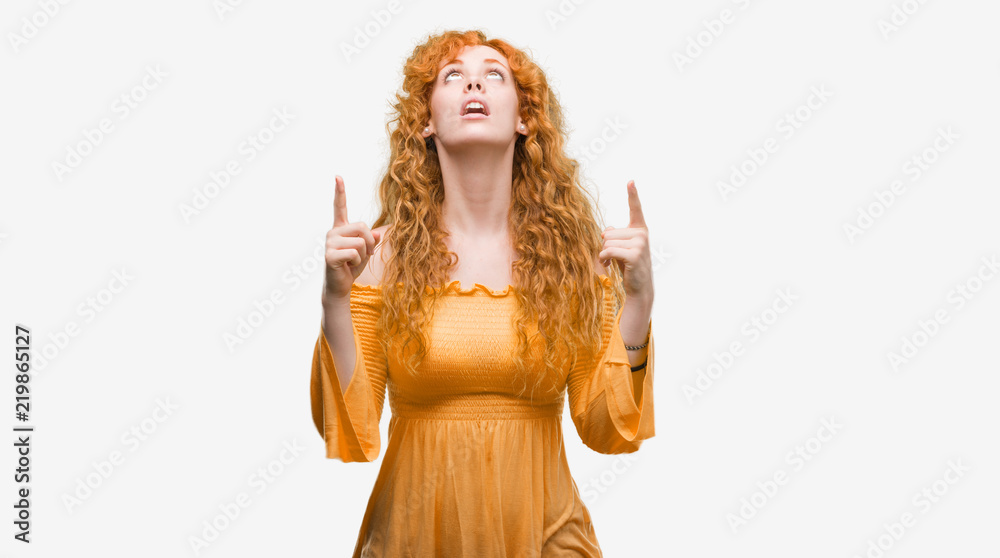 Young redhead woman amazed and surprised looking up and pointing with fingers and raised arms.