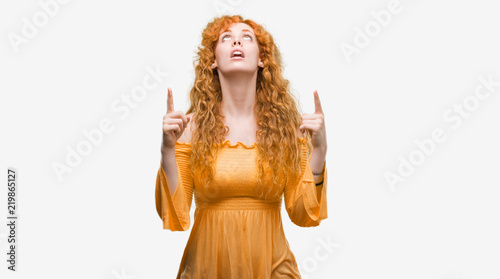 Young redhead woman amazed and surprised looking up and pointing with fingers and raised arms.