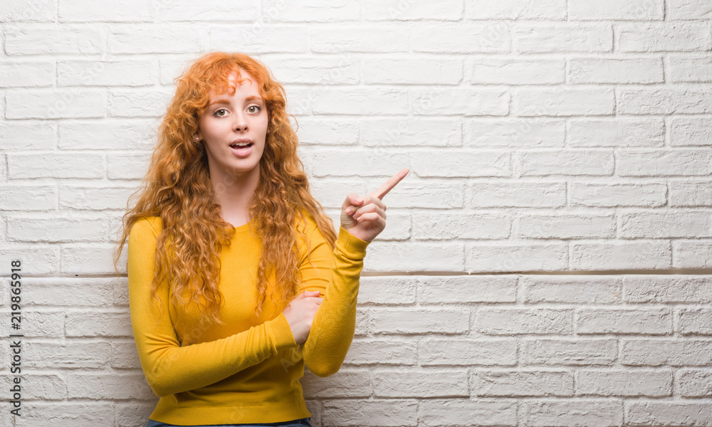 Young redhead woman standing over brick wall very happy pointing with hand and finger to the side Young redhead woman standing over brick wall very happy pointing with hand and finger to the side