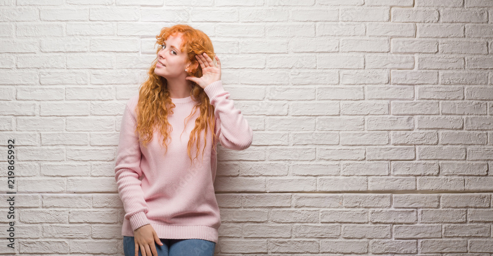 Young redhead woman standing over brick wall smiling with hand over ear listening an hearing to rumor or gossip. Deafness concept. Young redhead woman standing over brick wall smiling with hand over ear listening an hearing to rumor or gossip. Deafness concept.