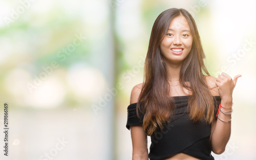 Young asian woman over isolated background smiling with happy face looking and pointing to the side with thumb up. © Krakenimages.com