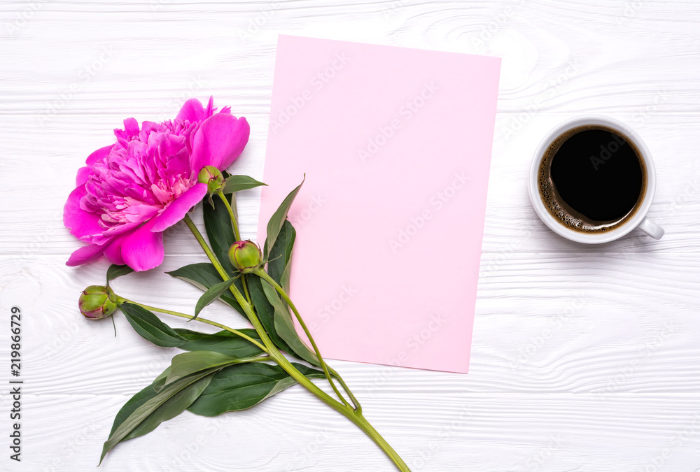 Empty paper with place for text, a cup of coffee and peony flower on a white wooden background. Mockup for your design. View from above.