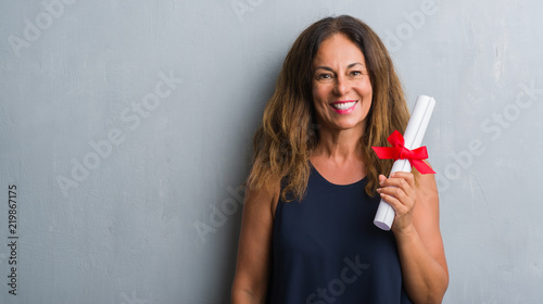 Middle age hispanic woman standing over grey grunge wall holding diploma with a happy face standing and smiling with a confident smile showing teeth