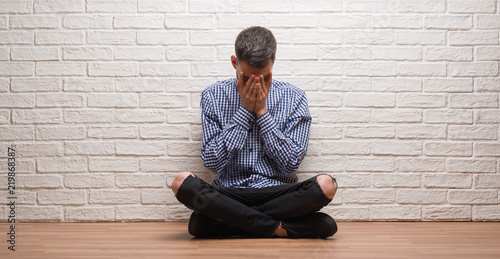 Young adult man sitting over white brick wall with sad expression covering face with hands while crying. Depression concept.