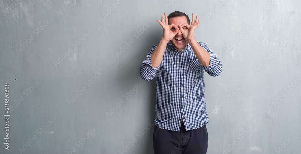 Young caucasian man over grey grunge wall doing ok gesture like binoculars sticking tongue out, eyes looking through fingers. Crazy expression.