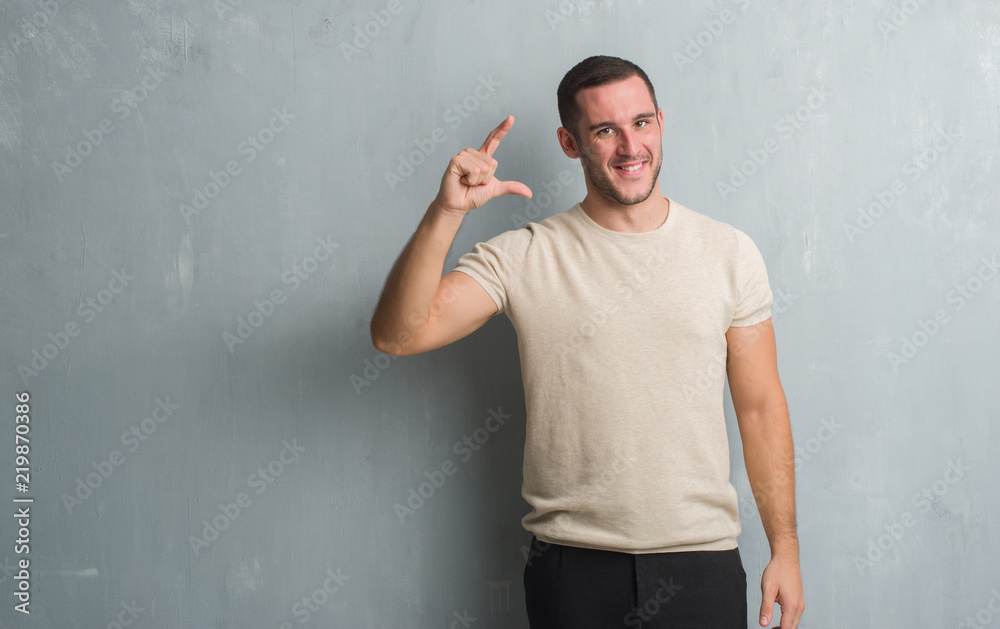 Young caucasian man over grey grunge wall smiling and confident gesturing with hand doing size sign with fingers while looking and the camera. Measure concept.