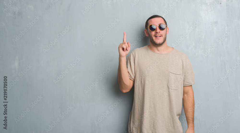 Young caucasian man over grey grunge wall wearing sunglasses pointing finger up with successful idea. Exited and happy. Number one.