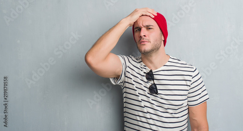 Young caucasian man over grey grunge wall wearing wool cap stressed with hand on head, shocked with shame and surprise face, angry and frustrated. Fear and upset for mistake.