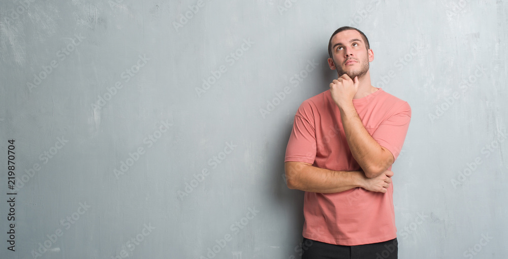 Young caucasian man over grey grunge wall with hand on chin thinking about question, pensive expression. Smiling with thoughtful face. Doubt concept.