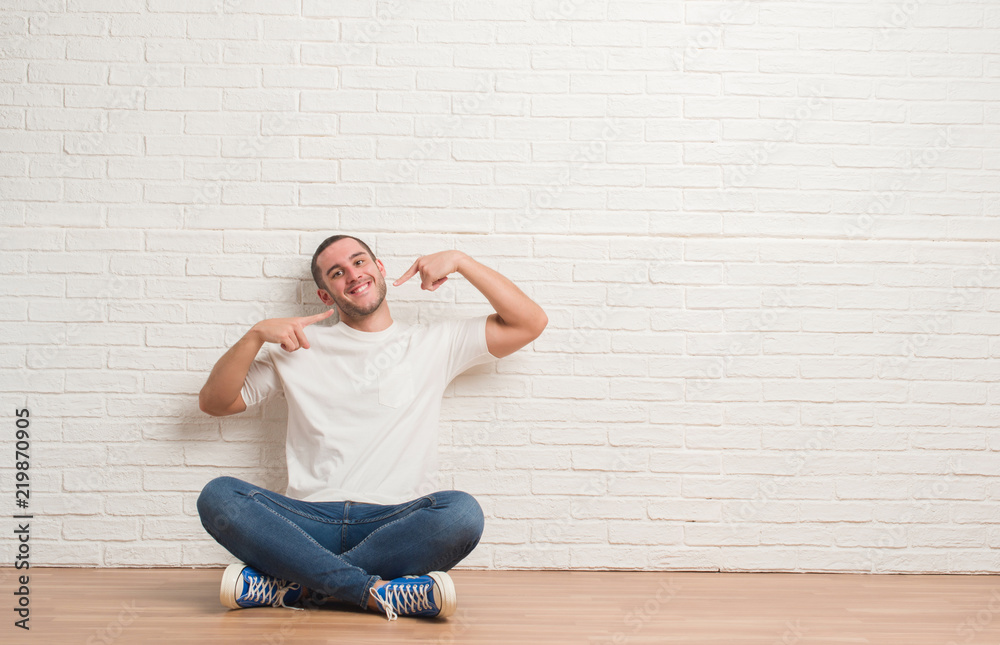 Young caucasian man sitting on the floor over white brick wall smiling confident showing and pointing with fingers teeth and mouth. Health concept.