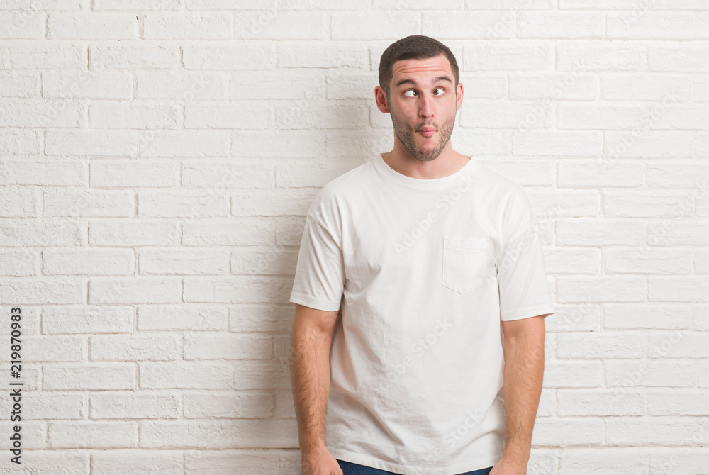 Young caucasian man standing over white brick wall making fish face with lips, crazy and comical gesture. Funny expression. Young caucasian man standing over white brick wall making fish face with lips, crazy and comical gesture. Funny expression.