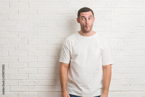 Young caucasian man standing over white brick wall making fish face with lips, crazy and comical gesture. Funny expression.
