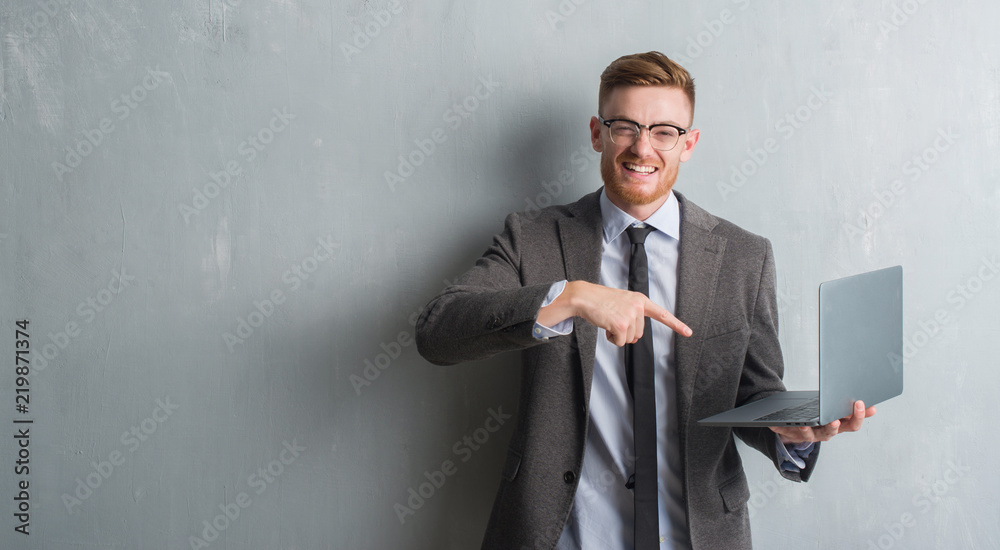 Young redhead elegant business man over grey grunge wall using laptop very happy pointing with hand and finger Young redhead elegant business man over grey grunge wall using laptop very happy pointing with hand and finger