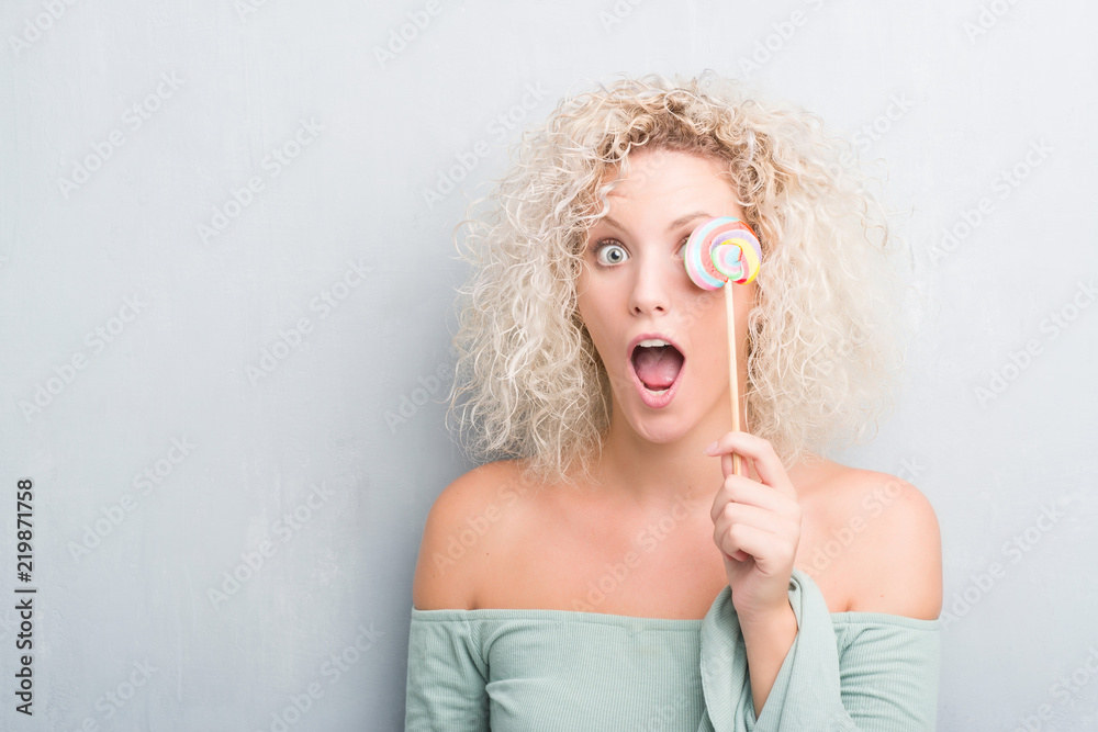 Young blonde woman over grunge grey wall eating candy lollipop scared in shock with a surprise face, afraid and excited with fear expression Young blonde woman over grunge grey wall eating candy lollipop scared in shock with a surprise face, afraid and excited with fear expression