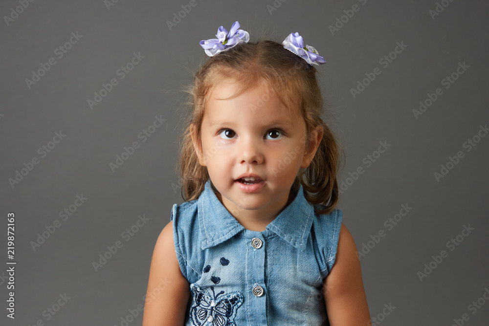 Happy face funny child girl in a blue dress. Portrait of adorable smiling little cute girl child with bow on her head isolated on gray background with copy space. Happy face funny child girl in a blue dress. Portrait of adorable smiling little cute girl child with bow on her head isolated on gray background with copy space.