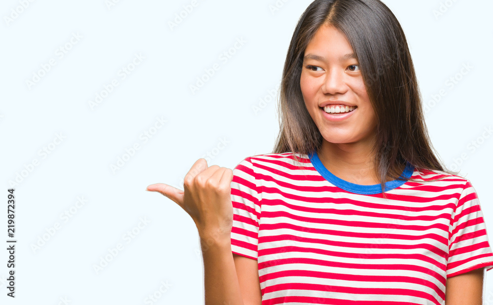 Young asian woman over isolated background smiling with happy face looking and pointing to the side with thumb up. Young asian woman over isolated background smiling with happy face looking and pointing to the side with thumb up.