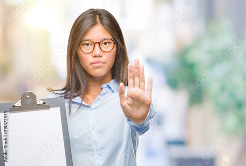 Young asian business woman over isolated background holding clipboard with open hand doing stop sign with serious and confident expression, defense gesture
