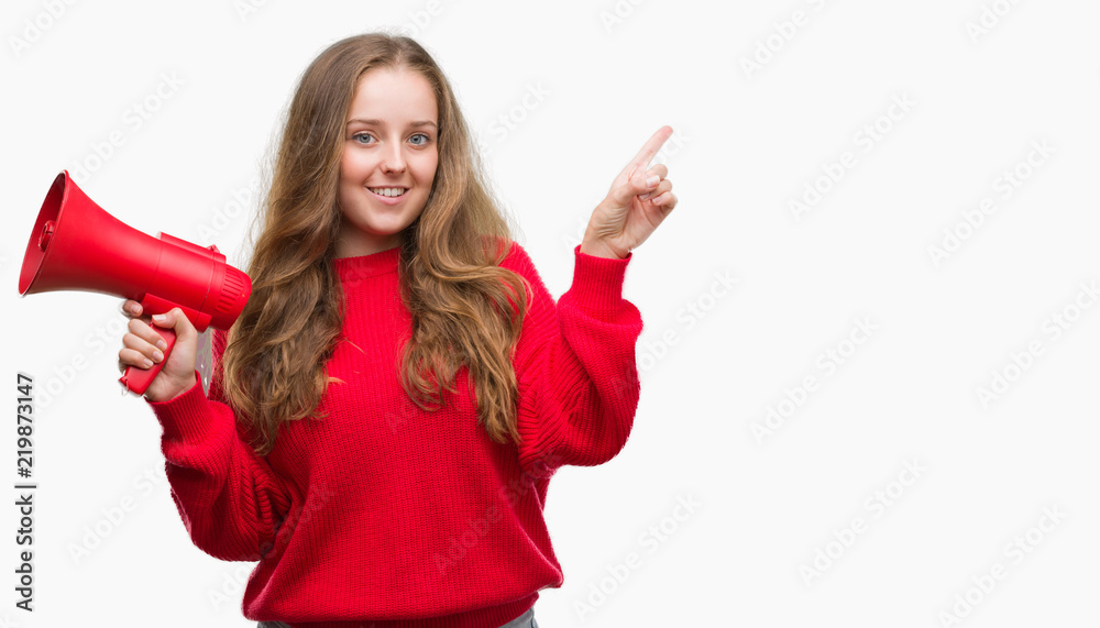 Young blonde woman holding red megaphone very happy pointing with hand and finger to the side Young blonde woman holding red megaphone very happy pointing with hand and finger to the side