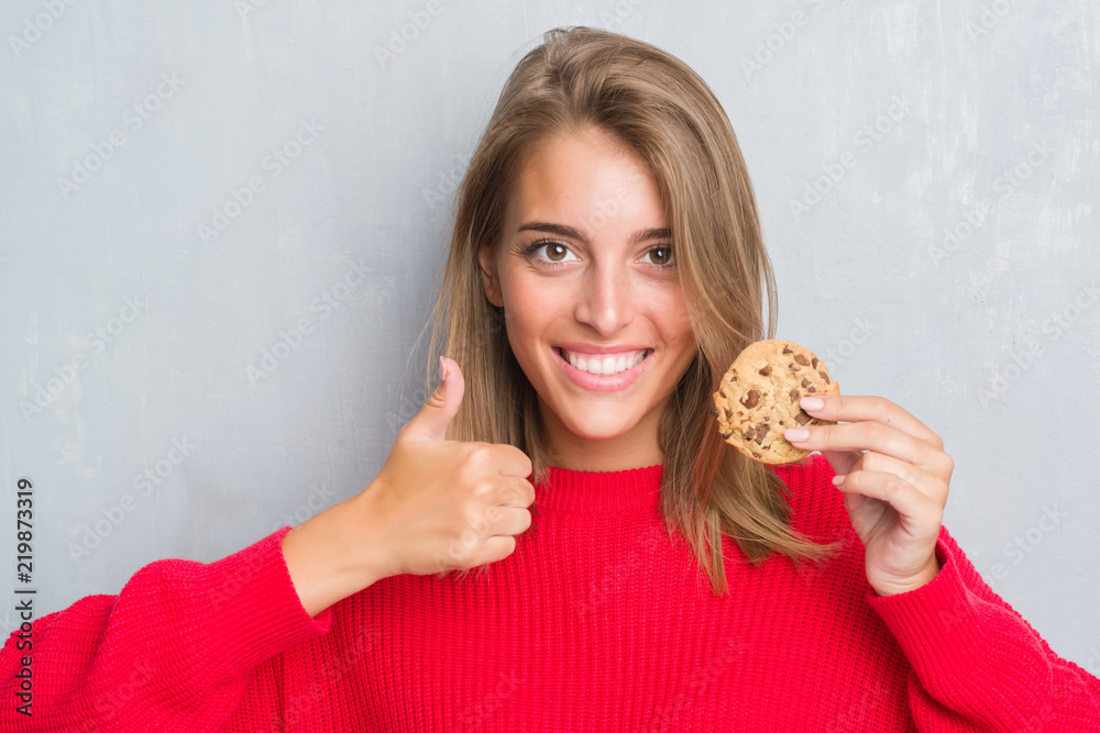 Beautiful young woman over grunge grey wall eating chocolate chip cooky happy with big smile doing ok sign, thumb up with fingers, excellent sign Beautiful young woman over grunge grey wall eating chocolate chip cooky happy with big smile doing ok sign, thumb up with fingers, excellent sign
